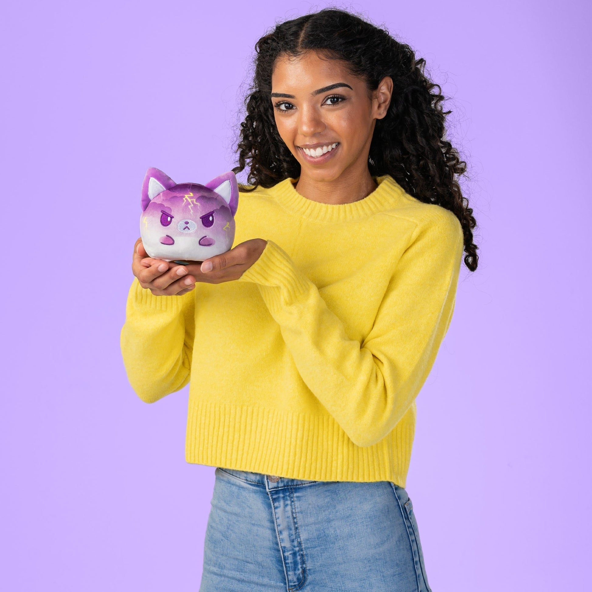 A young woman holding a Plushiverse Rain or Shine Fox 4” Reversible Plushie piggy bank on a plush background by TeeTurtle.