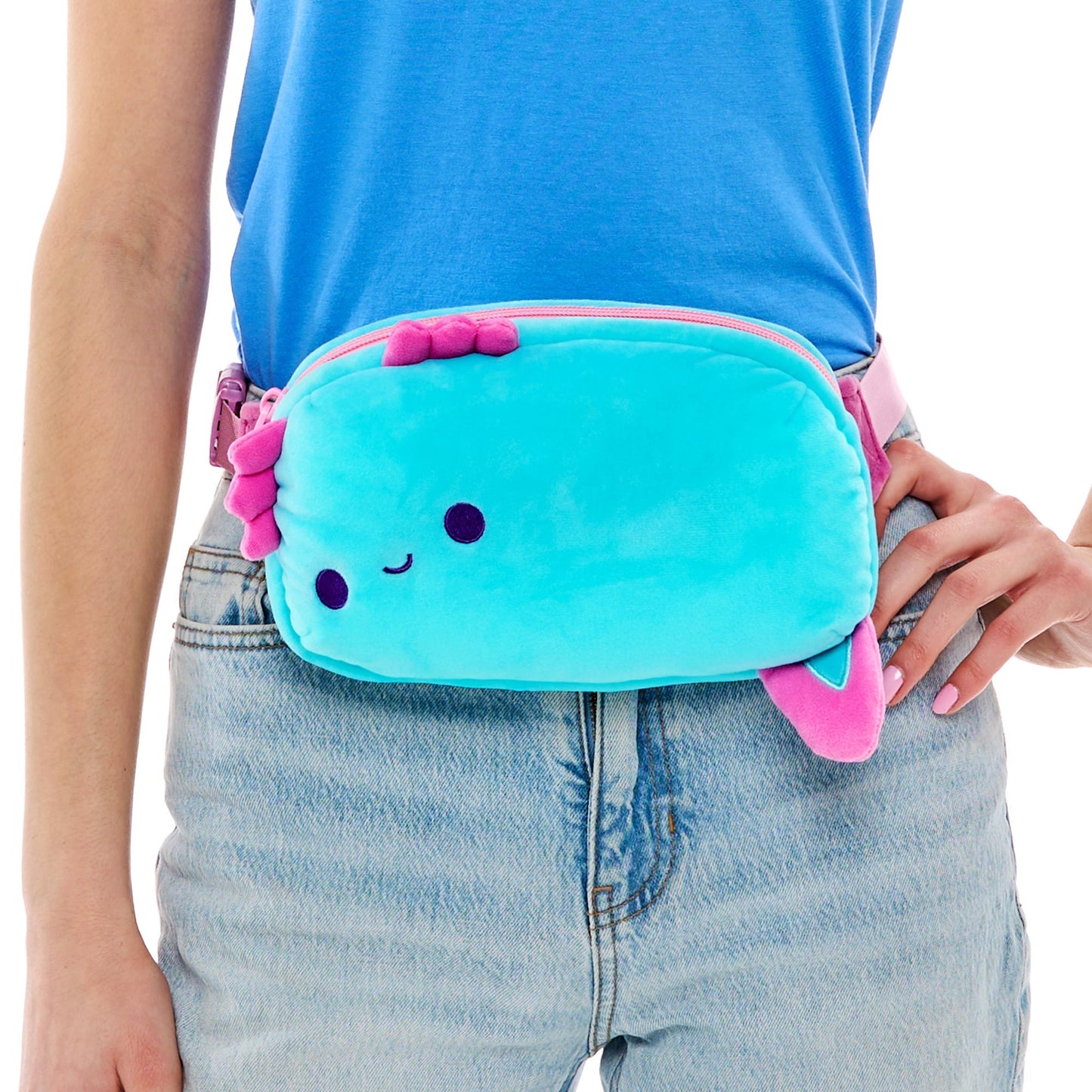 A person wearing jeans and a blue t-shirt is holding a cute, turquoise Plushiverse Alotl Fun Plushie Fanny Pack by TeeTurtle with a smiling face, pink accents, and an adjustable belt.