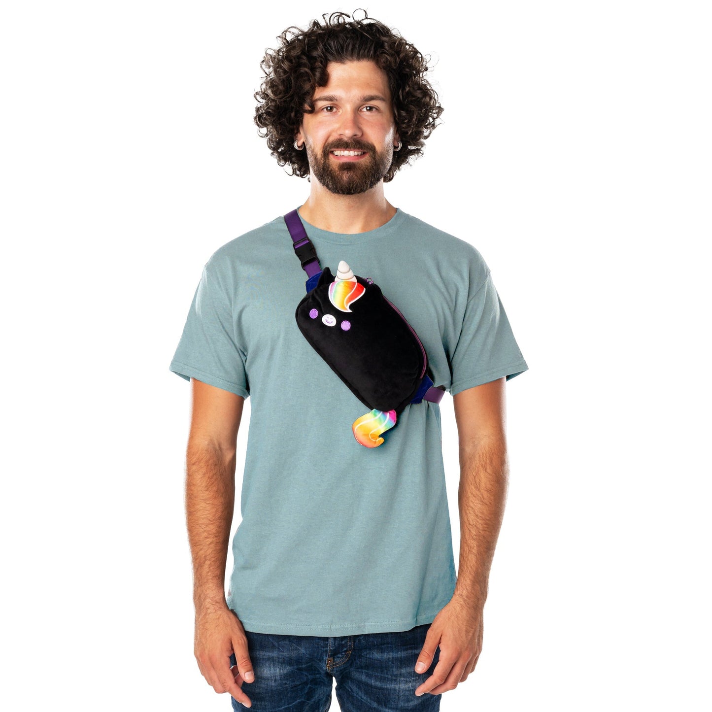 A man with curly hair and a beard is smiling and standing in a teal shirt. He is wearing a TeeTurtle Plushiverse Unicorn Pride Plushie Fanny Pack, showcasing hands-free functionality.