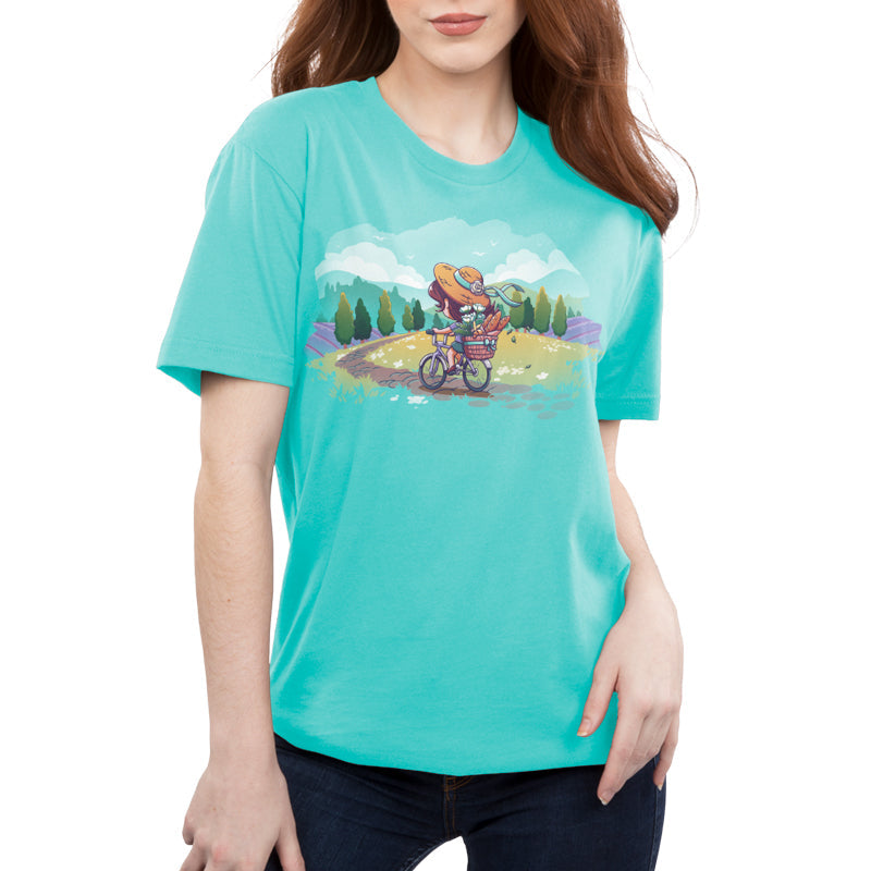 Premium Cotton T-shirt_TeeTurtle Countryside Biking Caribbean blue t-shirt featuring an illustration of a girl biking on a path in the countryside with bread and flowers in her basket. The path winds through rolling hills and fields of grass with small, white flowers. Green trees line the sides of the path, and lavender bushes are behind the trees, set against a light teal/turquoise sky, with fluffy white clouds and some birds.