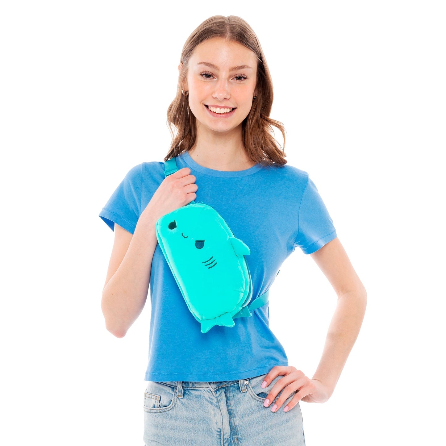 A young woman in a blue t-shirt and jeans smiles and holds a TeeTurtle Plushiverse Jaw-some Shark Plushie Fanny Pack.