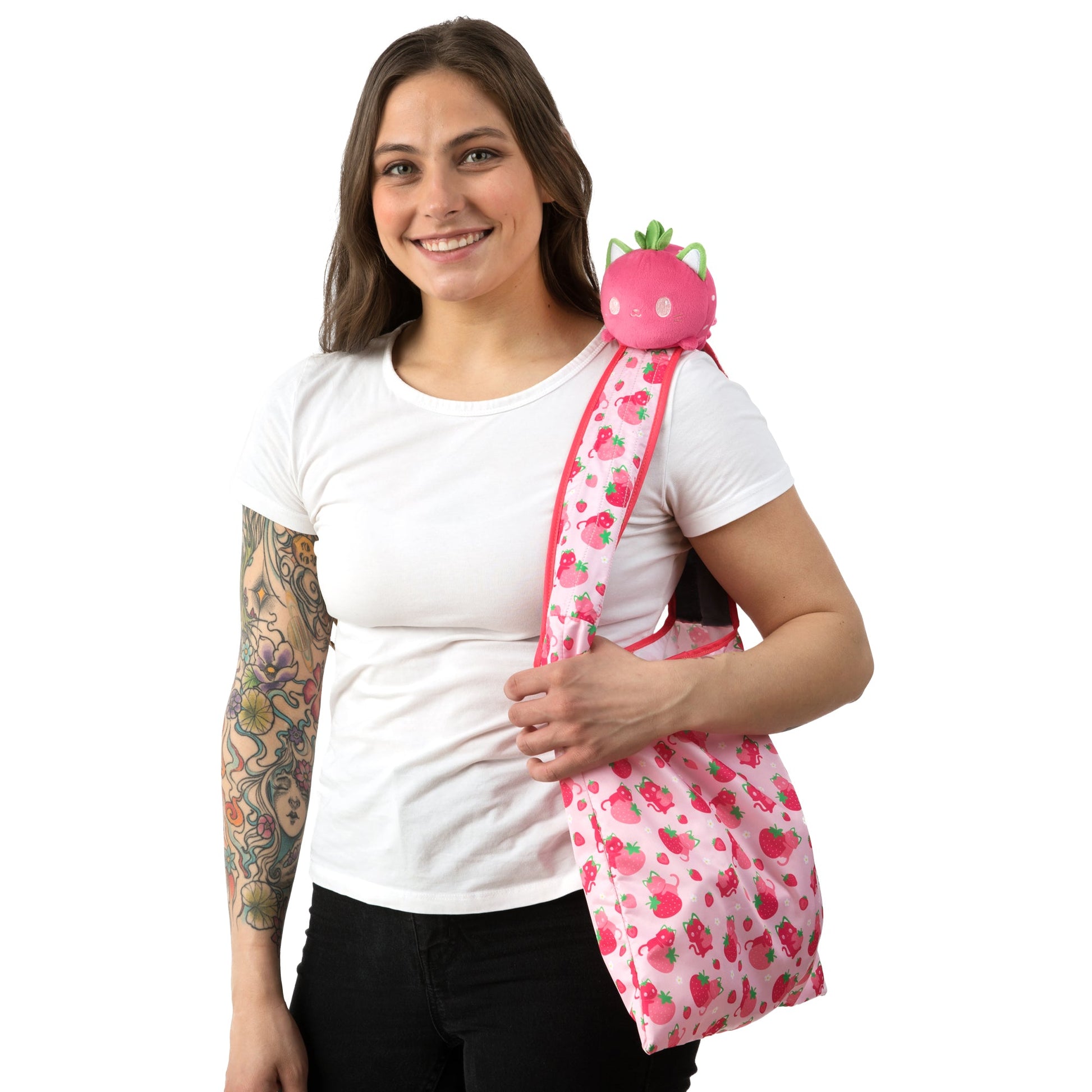 A woman with tattoos holding a pink Strawberry Cat Plushie Tote Bag by TeeTurtle.