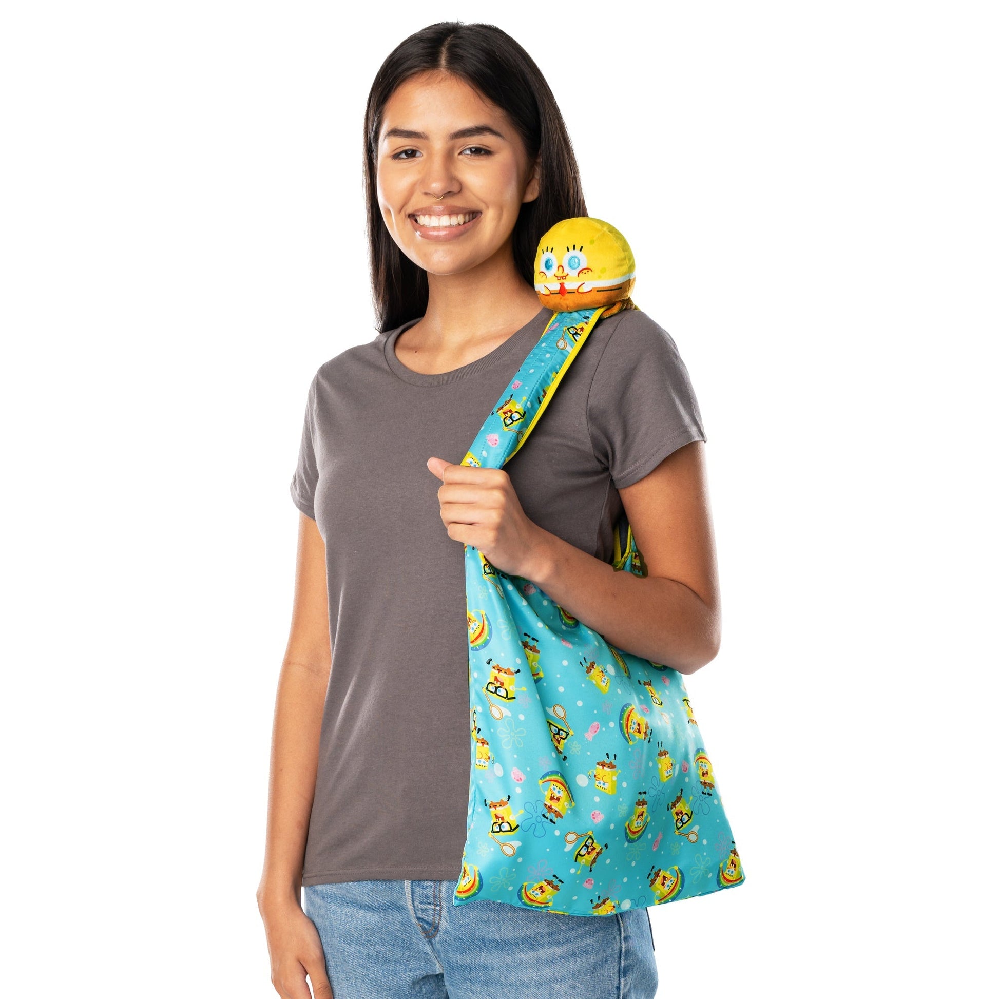 A woman wearing a gray t-shirt and jeans is smiling while holding a Plushiverse SpongeBob SquarePants Jellyfishing Plushie Tote Bag made from recycled plastic bottles, adorned with cartoon characters. She also has a Nickelodeon plushie on her shoulder.